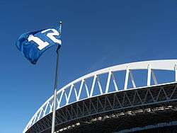 A blue flag with a white number 12 flies against a clear sky. An expansive white roof truss is behind the flagpole.