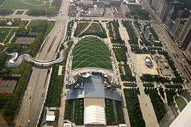  Aerial view of a green park with large roads running vertically at left and right and horizontally at the top. A curving metal bridge crosses the road on the left. Sidewalks divide the park into different areas, and it includes buildings and sculptures.