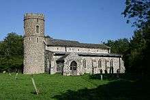 A flint church seen from the southeast; on the left is a round tower with a battlemented parapet, and the body of the church, with a porch and a small clerestory, extends to the right