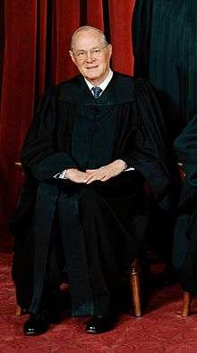 A balding old man with glasses, wearing a black robe and seated in front of a red curtain