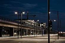 A night view of a viaduct running along the road. In the middle of the image is a glass station.