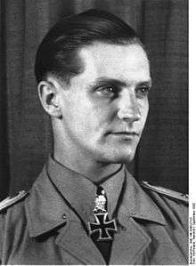 The head and shoulders of a young man, shown in semi-profile. He wears a military uniform with an Iron Cross displayed at the front of his shirt collar. His hair appears blond and short and combed back, his nose is long and straight, and his facial expression is determined but smiling; looking to the left of the camera.