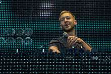 A picture of a man standing behind a turntable. He is holding a microphone in his hand