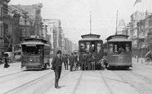 New Orleans, early years of 20th century. Canal Street, about halfway between the intersections of Magazine and Camp, looking lakewards along the neutral ground. Intersection with Camp Street visible at left; Chartres Street at right. Three electric streetcars are on the neutral ground, the route signs of two are visible, showing "Prytania" Street line and "Annunciation" Street line. A small group of men and boys standing around, including a police officer.