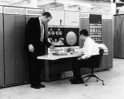 two men, one wearing glasses is seated, at a large computer