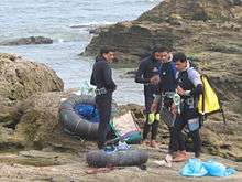  A croup of three divers dressed in wetsuits standing on a rocky shore with the sea in the background. On the ground are inflated truck inner tube floats with nets to support their catch