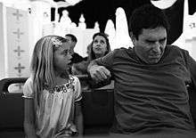 A black-and-white image of a young girl on the left and a middle-aged man sitting on a bench with a patterned background and others doing the same behind them. She is looking at him while he appears to grimace