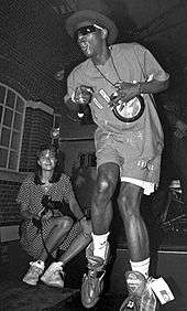 A man performing live onstage with a clock situated around his neck. A woman with a camera sits in the background.