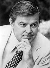 A man with salt-and-pepper hair, wearing a white jacket and shirt and polka-dotted tie, resting his chin on his left hand