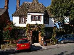 A front view of a two-storey public house, the lower storey in brick and the upper storey plastered.  It has a protruding central bay with a gable, steep tiled roofs and an ornate inn sign protruding diagonally from the right corner.