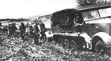 a black and white photograph of a half-tracked prime mover pulling heavy trucks along a muddy road