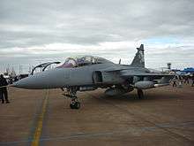 Three quarter view of grey jet fighter fitted with external fuel tanks on dirt-colored ground.