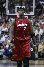 A man, wearing a red jersey with a word "MIAMI" and the number "7" written in the front, is walking on a basketball court.