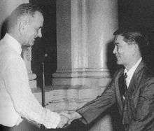 Tall Caucasian man standing in profile at left in a white suit and tie shakes hands with a smaller black-haired Asian man in a white shirt, dark suit and tie.