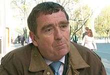 A man in a brown jacket, shirt and tie is pictured sitting outdoors in late spring