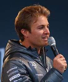 Man in his late twenties with a head full of hair and wearing a silver jacket. He is holding a microphone in his right hand.