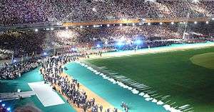 A portion of the stadium with stands full of people, a large artificial tree is on the right side of the image.  A group of people are walking together on the stadium floor