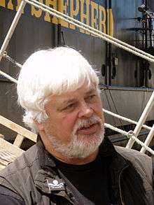 A heavyset man with white hair and a white beard, standing in front of a boat with the words "Shepherd" written on it in yellow paint