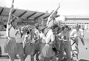 Women in hockey attire celebrate, waving their arms and sticks in the air