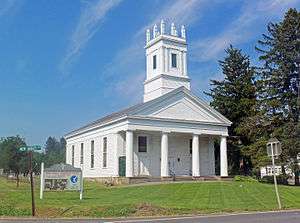 A white church with a colonnaded front and square steeple topped with spiky finials on the corners on a mowed grassy area under a blue sky. Behind it on the right are tall evergreen trees. In front on the left is a sign saying "Reformed Church of New Hurley" and a green street sign with "New Hurley Road" on it