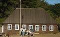 Photograph of the Fort Ross Commander's House on a sunny day. Visitors stand at a table in front of the rectangular log building with a high, peaked roof and white painted windowframes.