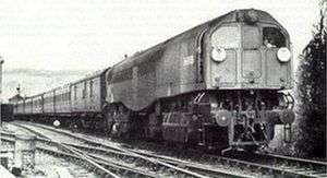 A side-and-front view of an 0-6-0+0-6-0 tank steam locomotive travelling towards the cameraman. The locomotive is a cuboid that contains boiler, fuel and cabs. The cuboid sits on two 0-6-0 bogies. It is hauling a train of around seven carriages past a signal box in the background.