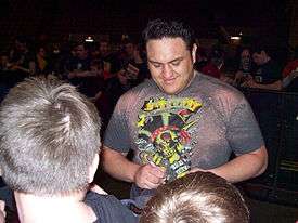 A dark-skinned adult male wearing a multi-colored T-shirt signing an autograph