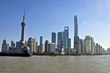 A group of skyscrapers and towers, seen from across a river. At the left is one consisting of a sphere on concrete supports topped by a long spike; in the center are smaller buildings, one a bright gold color, gradually rising to the tallest one at right, still under construction