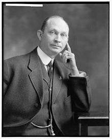 A balding man wearing a black jacket, vest, and tie, and white shirt, seated, leaning his left elbow on a table and his face against his extended left index finger