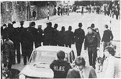 A line of police in the foreground facing some demonstrators/rioters in the middle distance in a city street.