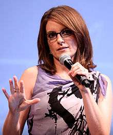 Tina Fey, a caucasian brown hair woman in her early 40s, wears a light purple paint splatter print dress. She wears dark-framed glasses, holds a microphone in her left hand, raises her right hand, and is in front of a light blue background.