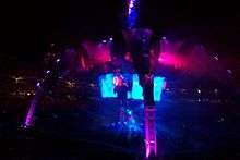 At night in a large outdoor stadium filled with people, a structure with four legs curving upward and meeting in a central spire is bathed in indigo and violet light, as a large circular video screen below the spire but above a stage shows a dark-clothed man with arm raised singing into an illuminated circular microphone.