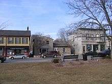 A row of three older buildings lines the far side of a street. On the near side is grass from a park with two park benches