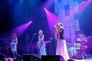 Four female musicians performing live on-stage against a black and white backdrop with violet lights.