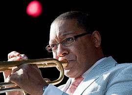 An African-American male playing a trumpet. He is wearing glasses and a light blue suit.