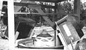 A sphere in s cyclider in the ground, above which is wooden scaffolding and two large boxes. In the background are trees.